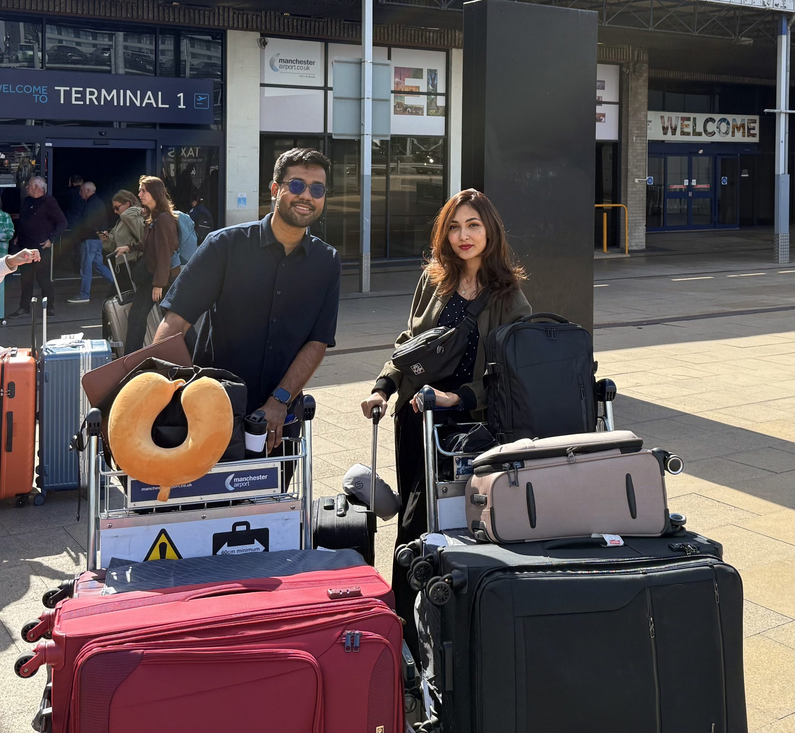 Two people stand outside an airport terminal with trolleys loaded with suitcases and bags, beginning their Chevening journey. A large neck pillow sits atop one trolley, with the terminal building and a "WELCOME" sign in the background.