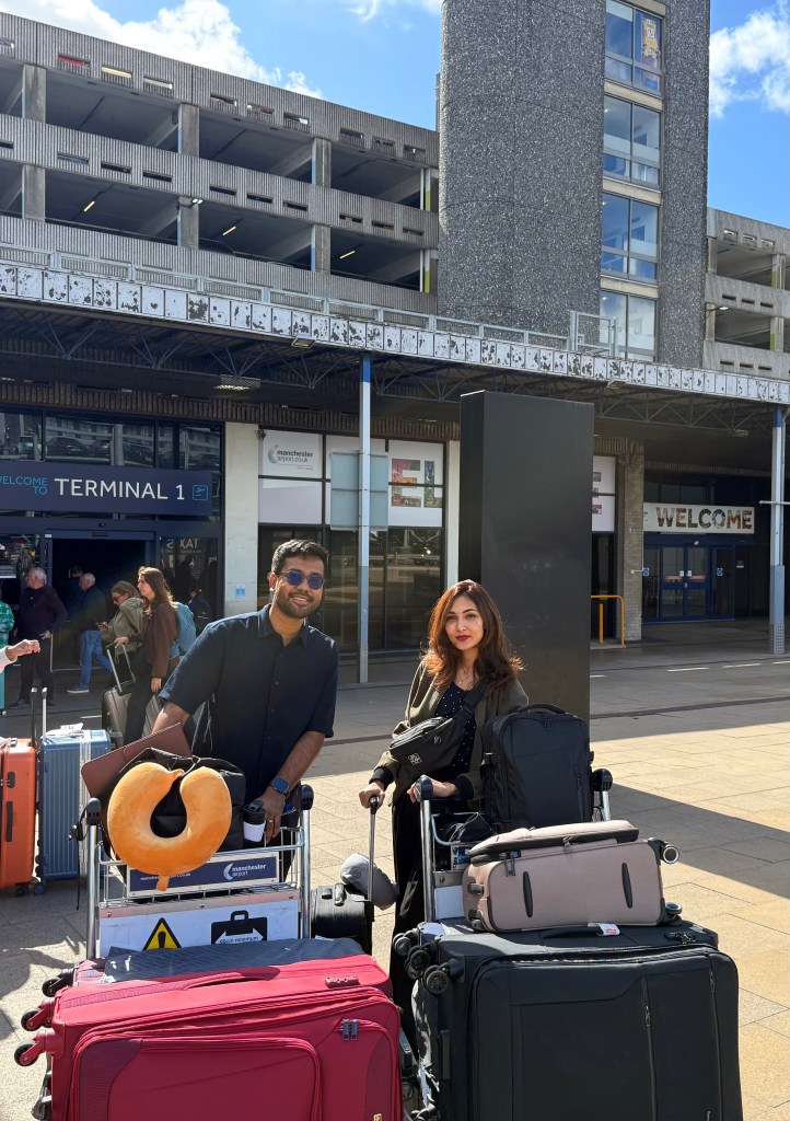 Two people stand outside an airport terminal with trolleys loaded with suitcases and bags, beginning their Chevening journey. A large neck pillow sits atop one trolley, with the terminal building and a "WELCOME" sign in the background.