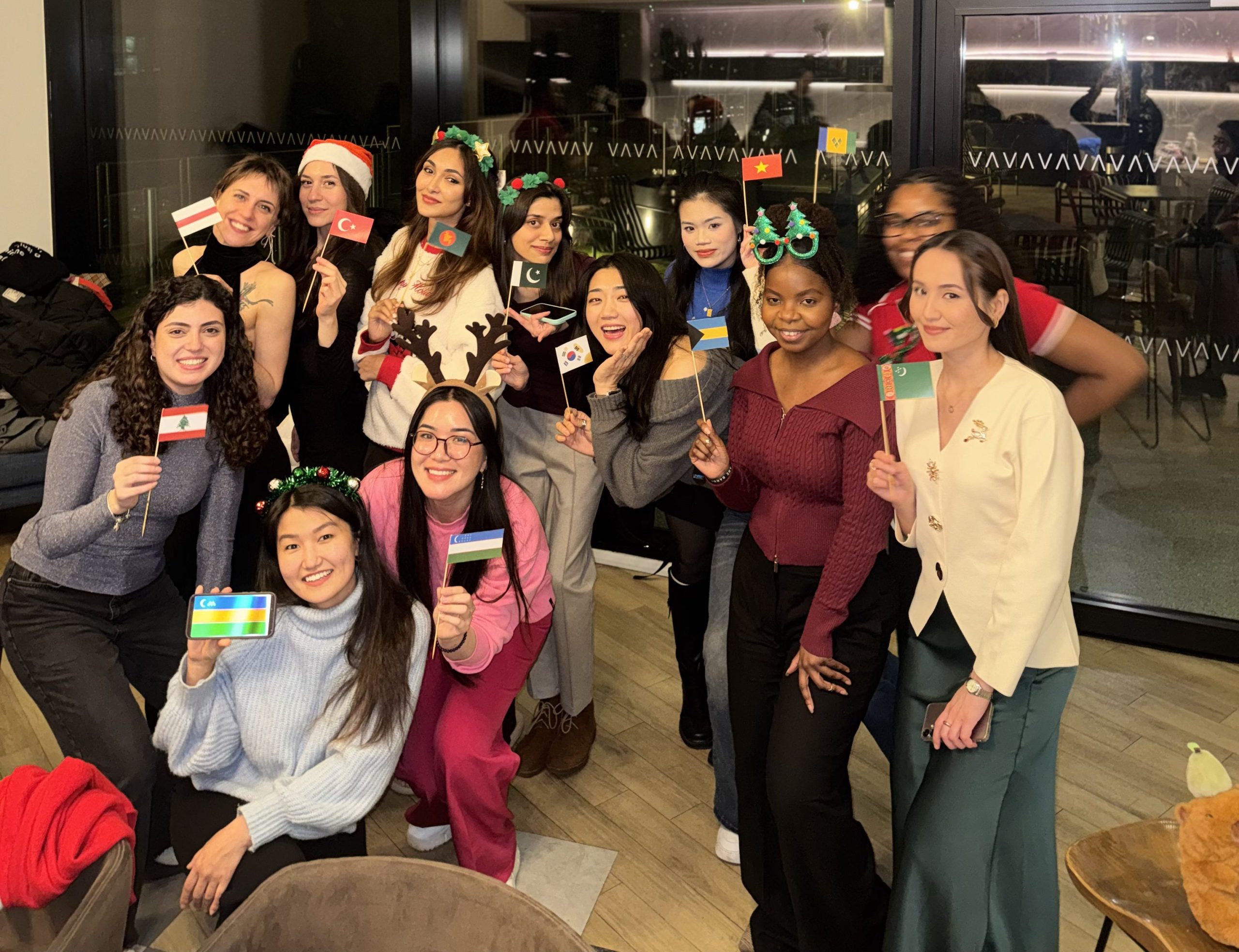 A group of women pose together indoors, many wearing festive headbands and holding small holiday-themed cards. Large windows behind them reveal city lights and tall buildings at night, capturing a joyful moment in their Chevening journey.
