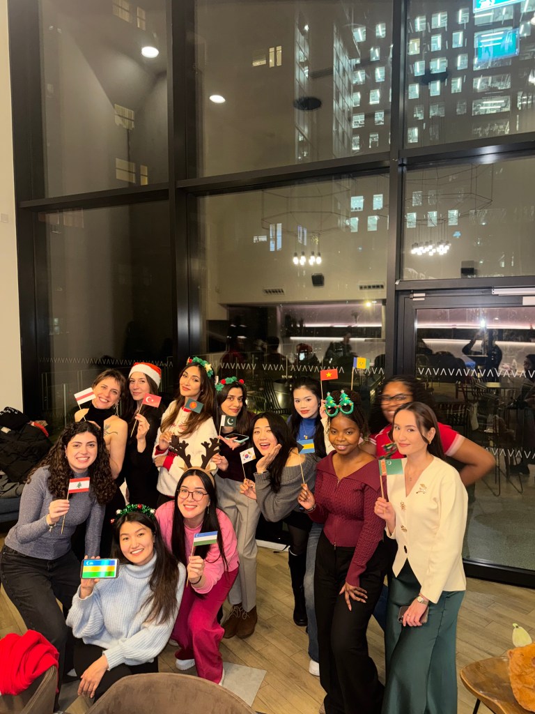 A group of women pose together indoors, many wearing festive headbands and holding small holiday-themed cards. Large windows behind them reveal city lights and tall buildings at night, capturing a joyful moment in their Chevening journey.