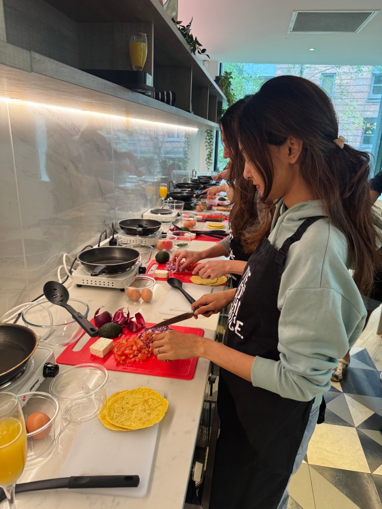 Two people wearing black aprons are standing at a kitchen counter, chopping vegetables on red cutting boards. Their teamwork and shared laughter could easily be part of inspiring student stories on a Chevening journey in this modern kitchen with shelves and potted plants above.