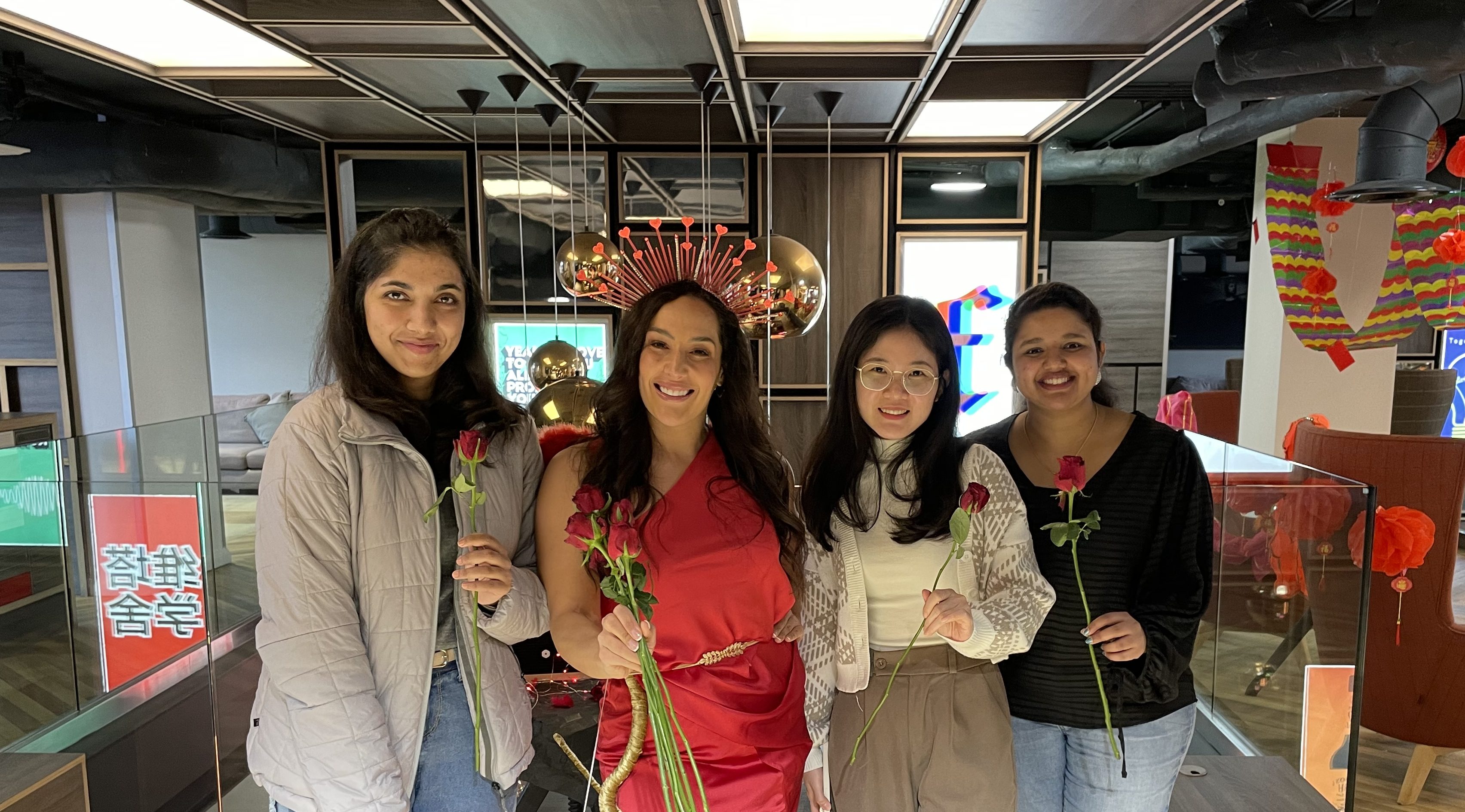 Four women stand indoors, smiling and holding red roses. One wears a bright red dress and a decorative headpiece, while the others are in casual clothing. Festive decorations set the scene for memorable Vita Student stories.