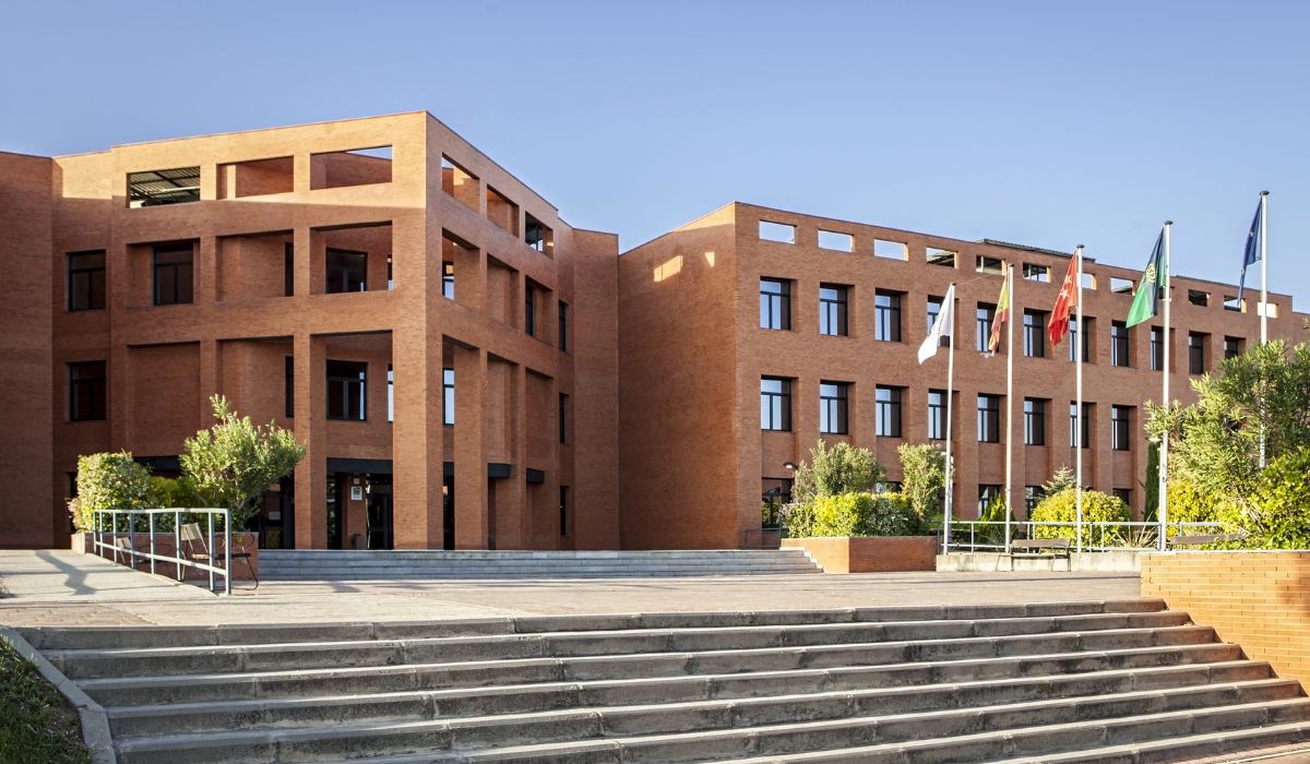 A modern, angular brick building with large windows offers stylish student accommodation in Madrid. Surrounded by greenery, it features flagpoles out front and wide concrete steps leading to the entrance under a clear blue sky.