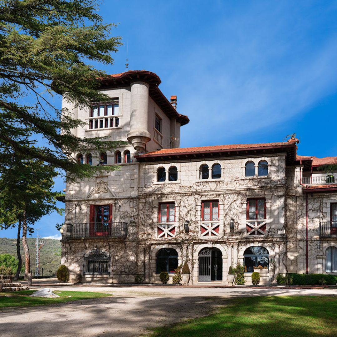 A large stone building offering student accommodation in Madrid features red-framed windows, arched doorways, and a central tower. Surrounded by trees under a clear blue sky, shadows fall gracefully across the facade and lawn.