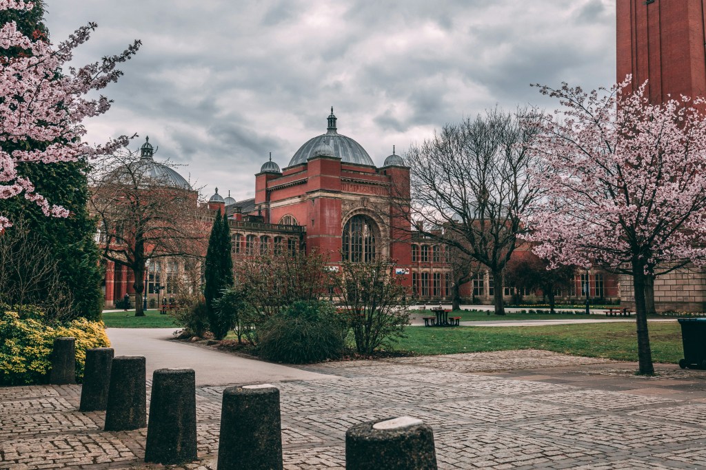A red-brick university building with domed roofs, recognized in the QS World University Rankings 2027, stands behind a green lawn and blossoming cherry trees under a cloudy sky, viewed from a cobblestone path lined with bollards.
