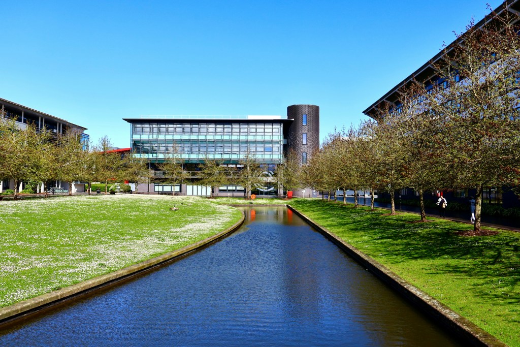 A modern glass and brick building stands by a narrow canal, flanked by grassy areas with trees and sidewalks, under a clear blue sky—home to an institution featured in the 2027 QS World University Rankings.