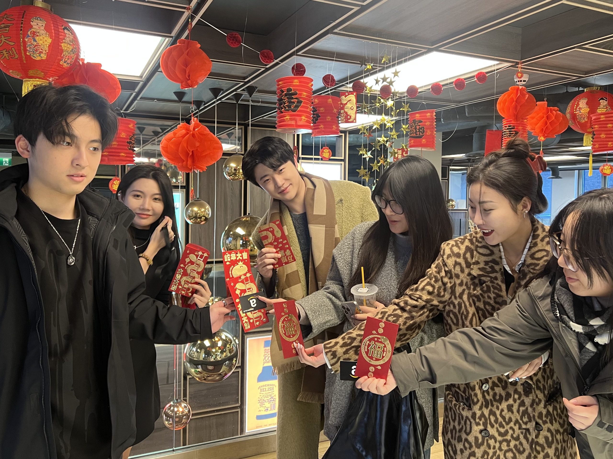 Six young people stand indoors decorated with red lanterns and festive ornaments, holding red envelopes and smiling together, each sharing their own idea of home as they celebrate a traditional festival.