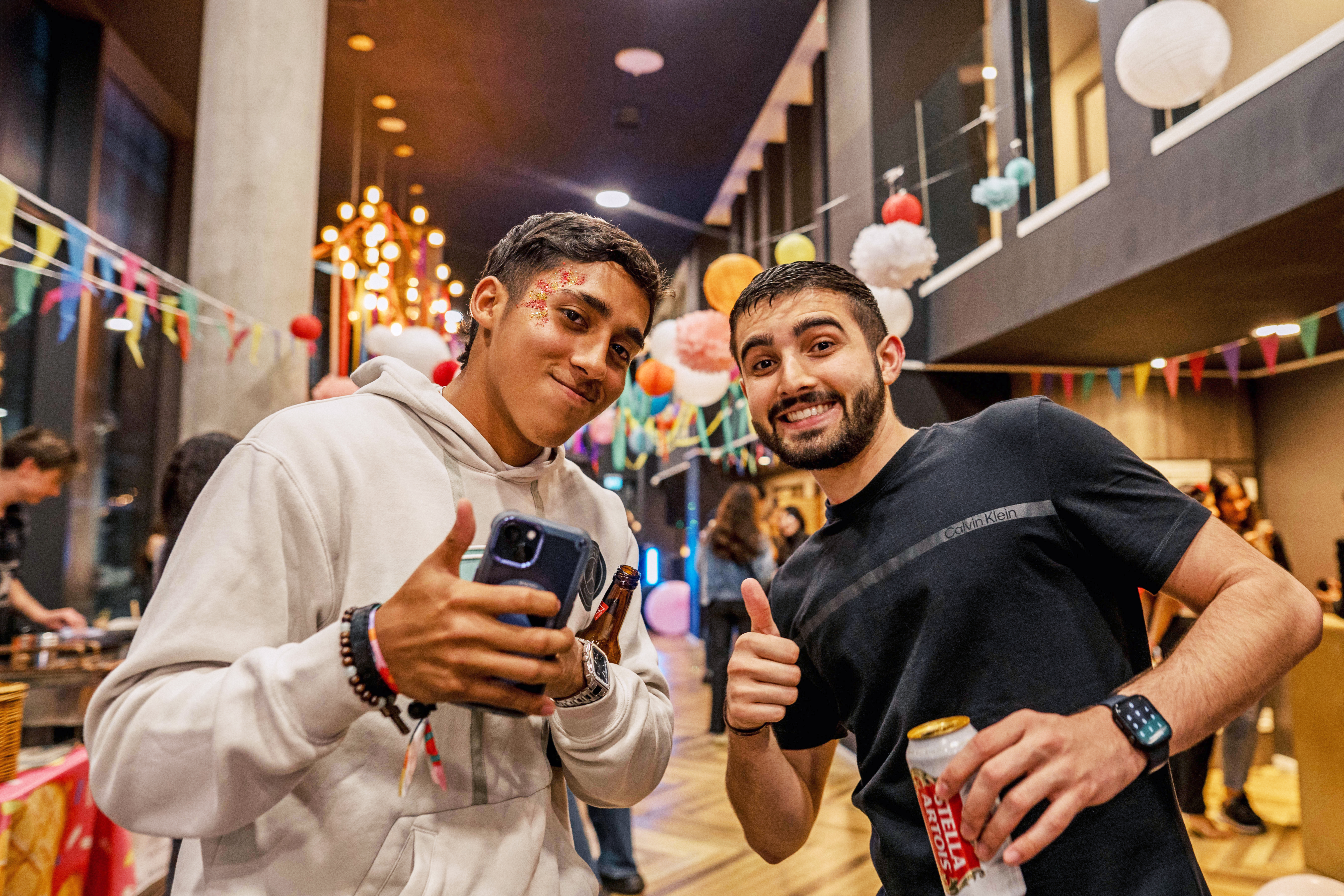 Two young men smile at a festive indoor Vita Student Spain party. One holds a phone and wears a white hoodie with glitter on his face; the other, in a dark t-shirt, gives a thumbs up and holds a drink. Colorful decorations hang in the background.
