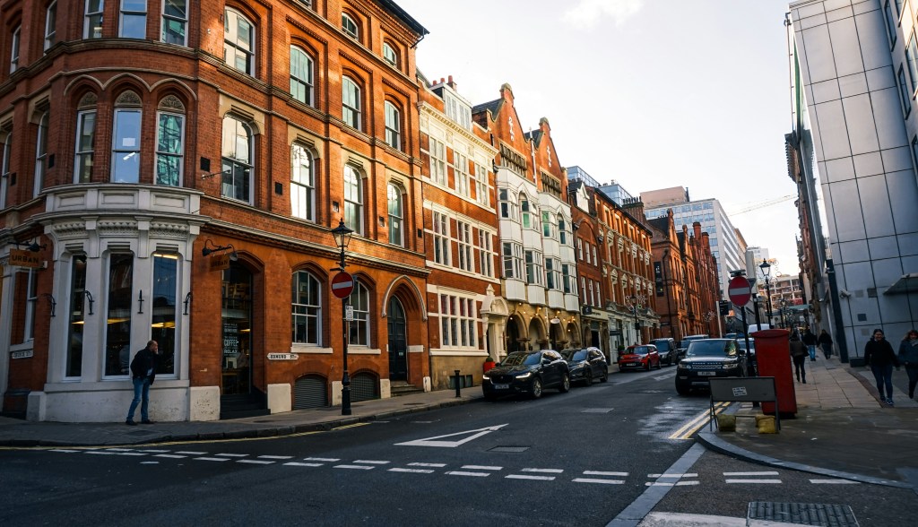 A city street, like Gough Street, lined with red-brick and white buildings, cars parked along the road, students and other pedestrians strolling the sidewalks, and a red mailbox on the corner under a partly cloudy sky.