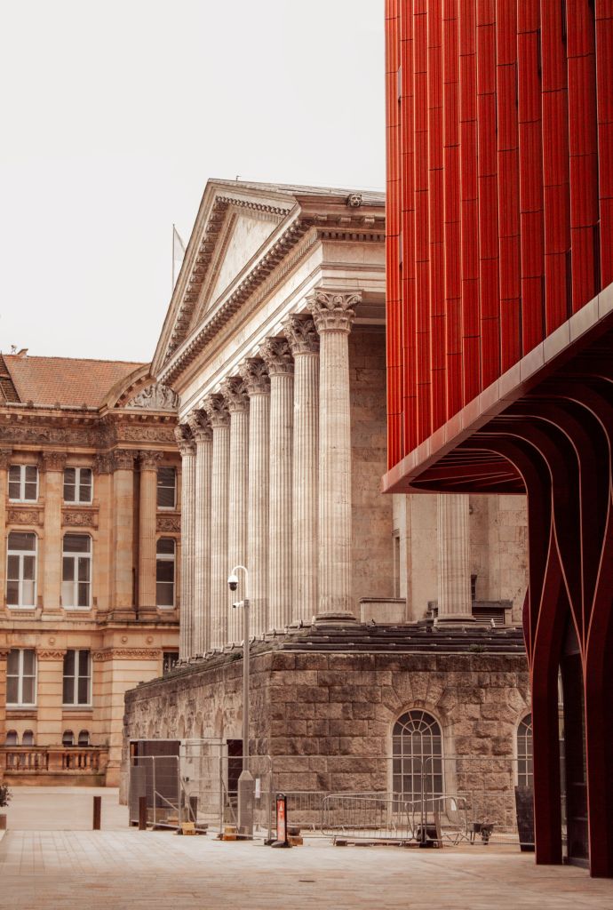 A neoclassical building with tall stone columns stands beside a modern structure with red vertical panels and curved supports on Gough Street in an urban setting.