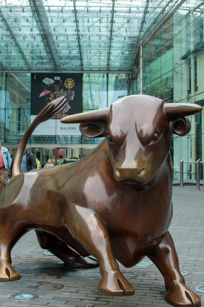 A large bronze bull statue stands on a paved area near Gough Street, in front of a glass building entrance, with people and students passing by and a colorful billboard visible in the background—one of the unique things to do nearby.