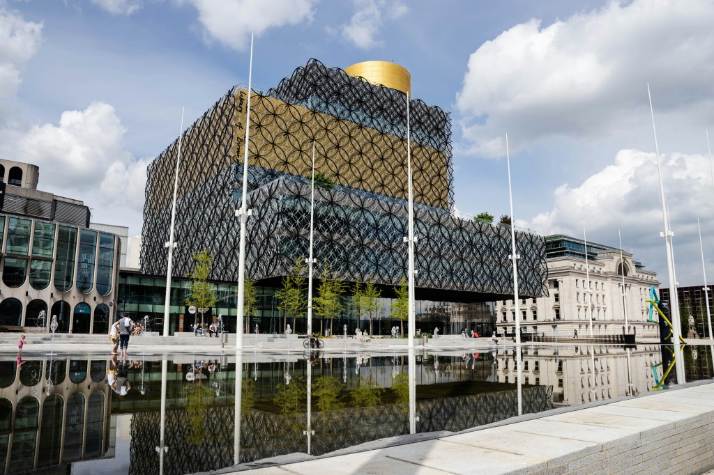 A modern building with a unique circular-patterned facade and a golden rooftop stands beside a reflective pool on Gough Street, with students and others walking nearby under a partly cloudy sky—an inviting spot for your list of things to do.