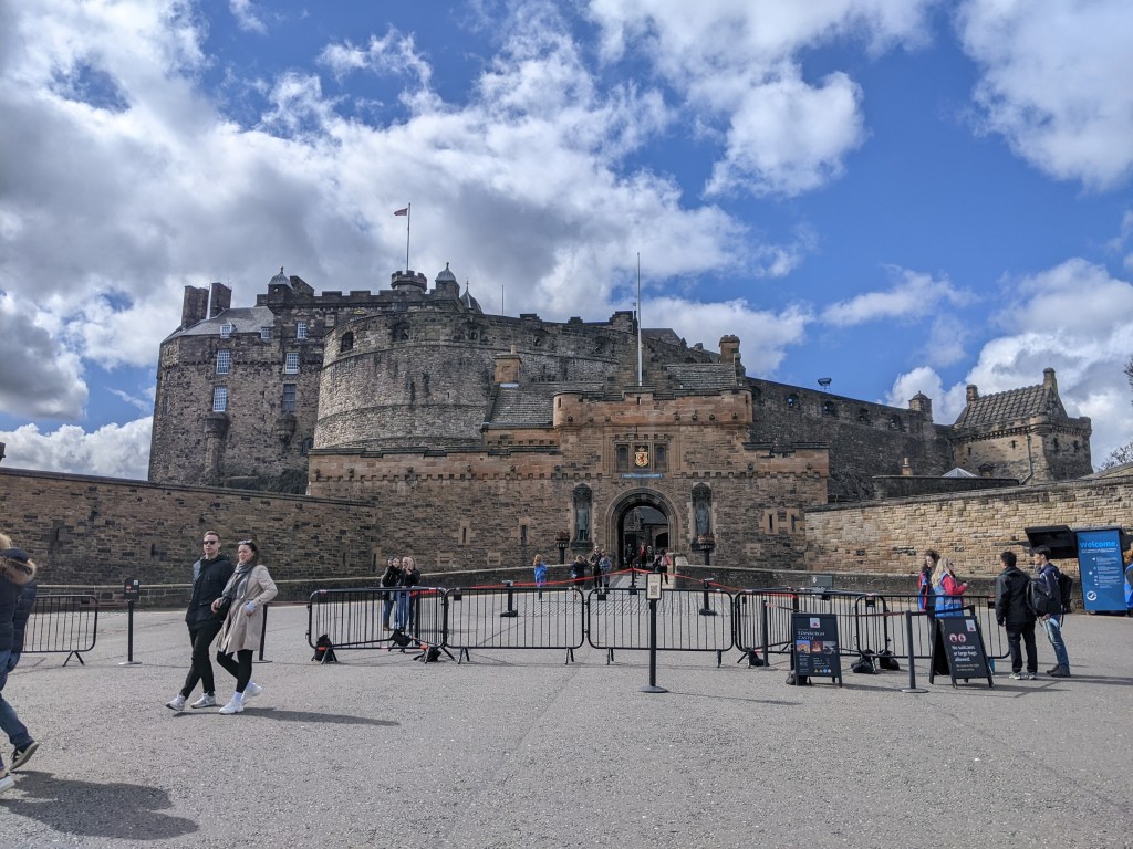 Edinburgh Castle stands atop a rocky hill under a partly cloudy sky, with visitors—including students who live in Edinburgh—gathering near the entrance, surrounded by barriers and informational signs at one of the city’s best places to visit.