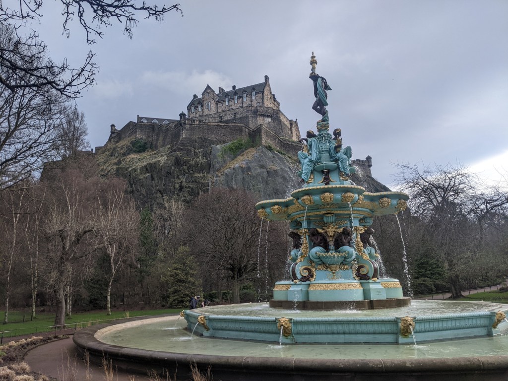 A colorful ornate fountain with statues stands in the foreground, while one of the best places to live in Edinburgh—a historic stone castle—perches on a rocky hill surrounded by trees under a cloudy sky.