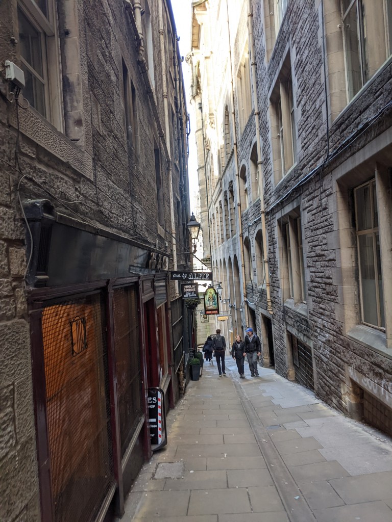 A narrow cobbled alleyway between old stone buildings, close to some of the best places to live in Edinburgh. Signs and lamps hang from the buildings as sunlight filters through, with several people walking in the distance.