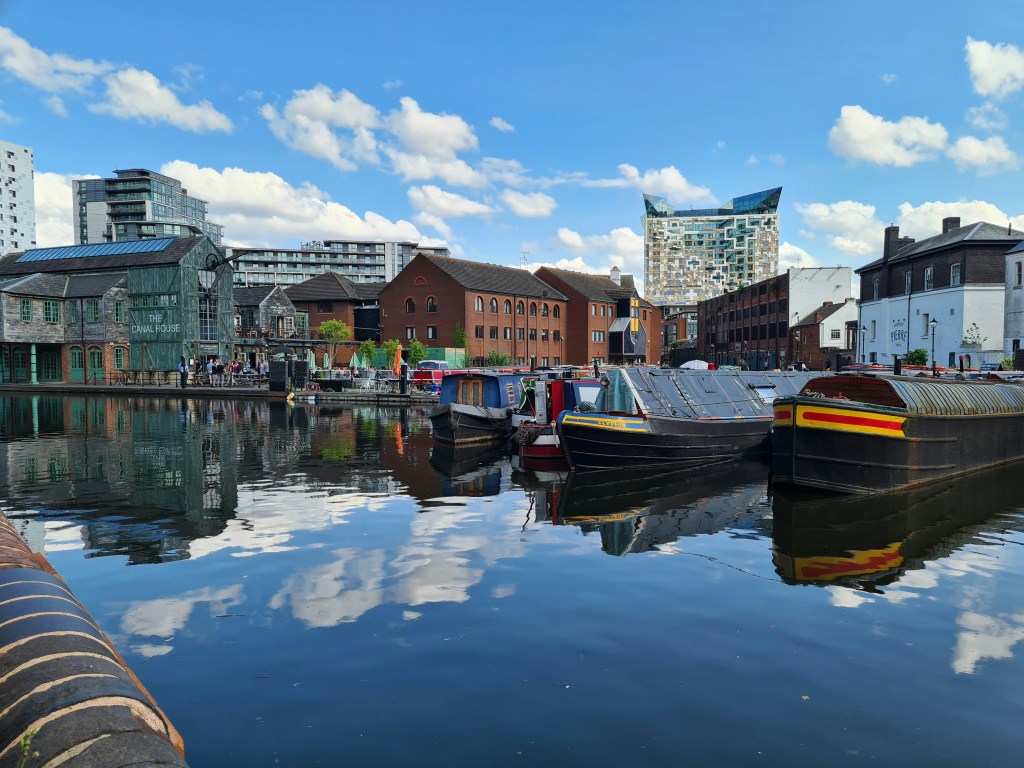 Canal boats are moored along a calm waterway near Gough Street, reflecting blue sky and clouds. Modern and older brick buildings line the banks, offering students plenty of things to do in this lively area, with a distinctive glass building visible in the background.