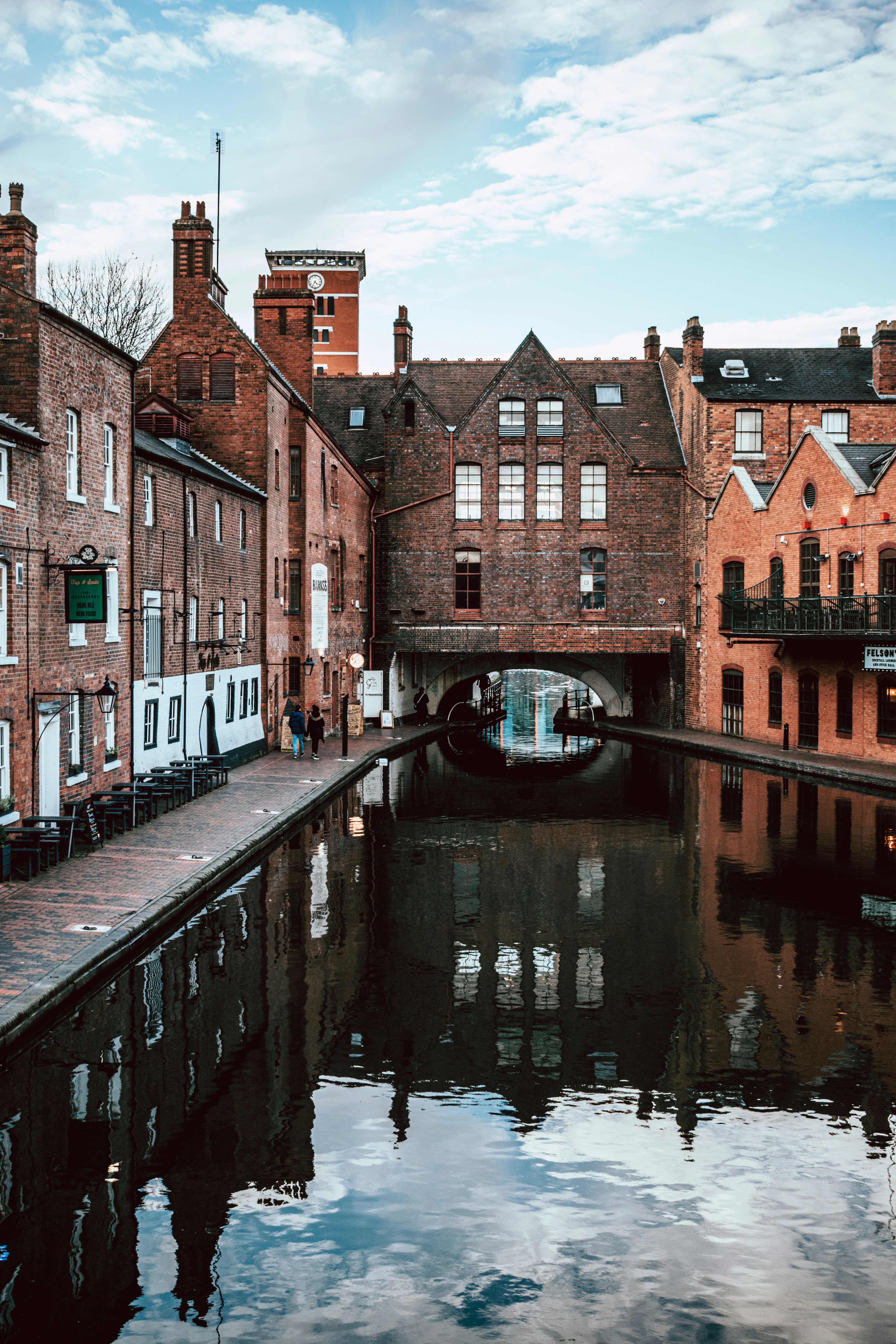 A canal runs alongside historic brick buildings under a partly cloudy sky on Gough Street. The buildings are reflected in the calm water as students and locals walk along the cobblestone path, exploring things to do by the water.
