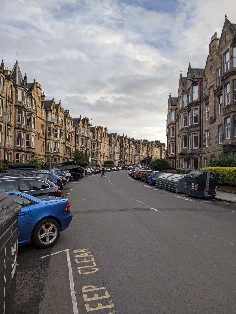 A curved street lined with parked cars and tall, brown stone buildings under a cloudy sky—classic scenery near some of the best student accommodation Edinburgh has to offer, with “KEEP CLEAR” painted on the road in the foreground.