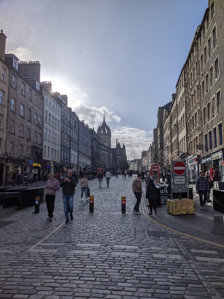 A cobblestone street lined with historic buildings, people and students walking, and the sun shining through clouds; a church with a tall spire is visible in the distance—one of the best places to live in Edinburgh.