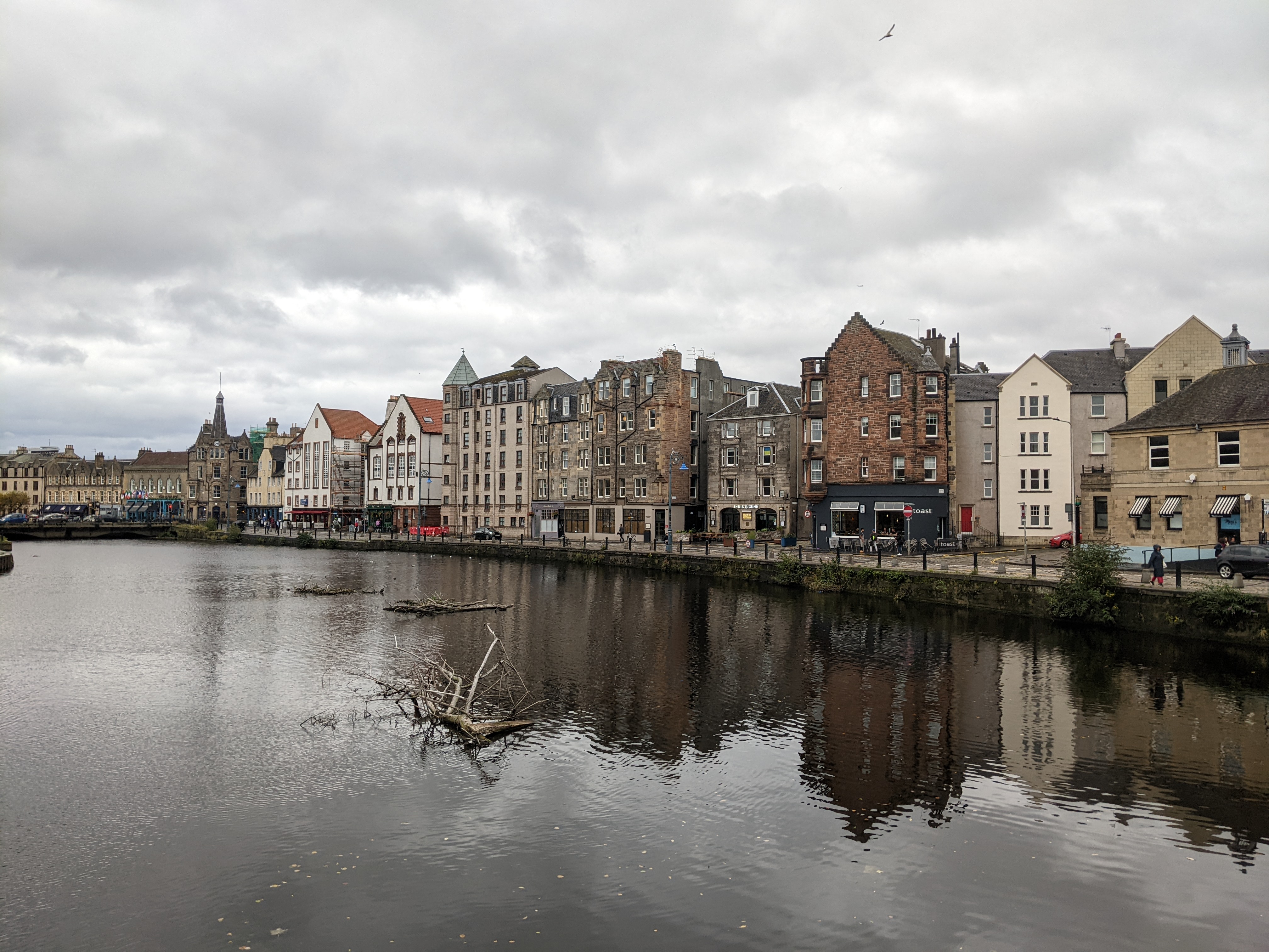 A row of colorful, historic buildings lines a waterfront under a cloudy sky—one of the best places to live in Edinburgh. The calm water reflects the scene, while a small tree branch floats in the river.