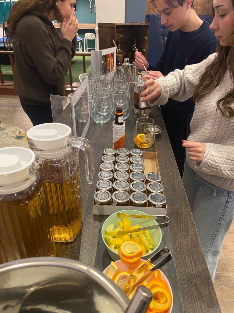 Three people stand at a counter in York preparing drinks with jars of spices, sliced citrus fruits, and pitchers of a golden beverage. Glasses and bottles are arranged on the counter, creating a casual and social Student Stories moment.