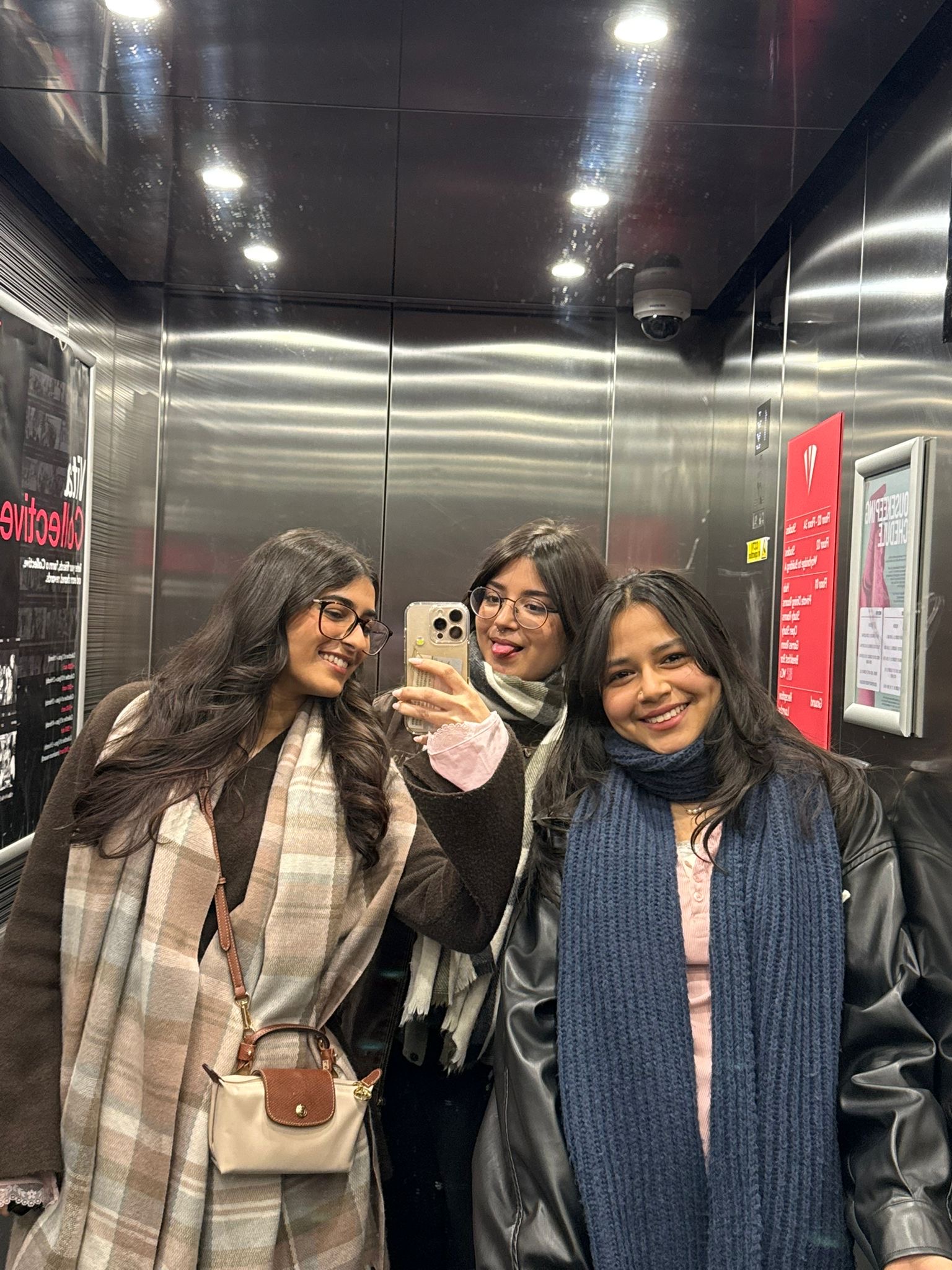 Three women smile and take a selfie together in an elevator, wearing winter clothing like shawls, scarves, and coats. The shiny metallic walls and visible posters hint at the vibrant atmosphere of Vita Student residences—a perfect backdrop for unforgettable student stories.