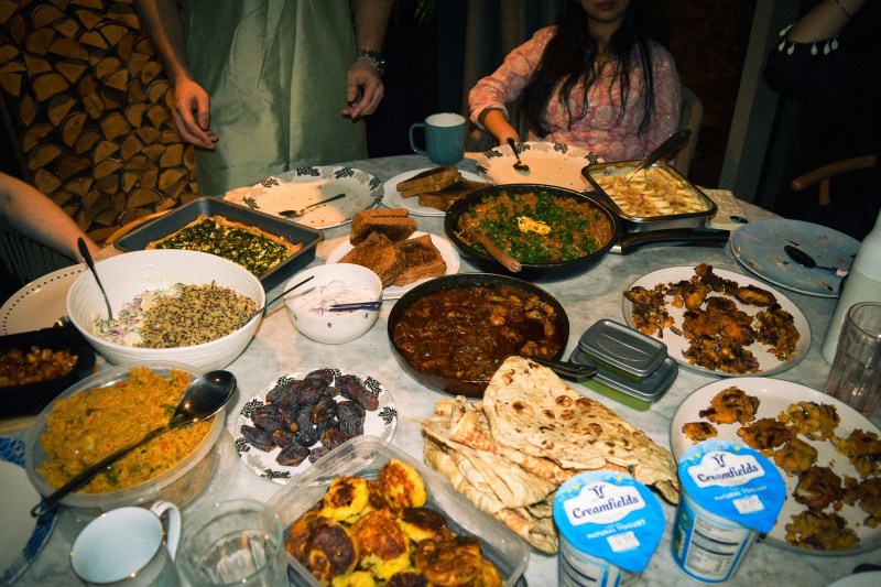 A dining table filled with various dishes, breads, and desserts reflects the warmth of Vita Student living. Four people sit or stand around the table, sharing student stories and preparing to eat. Multiple plates and cups are visible among the shared food items, capturing a sense of community that makes rebooking an easy choice each year.
