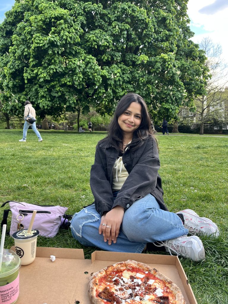 A woman sits cross-legged on grass in a park, smiling as she enjoys her lunch—a pizza box, drink, and bag are in front of her. Large green trees and people walking in the background set the scene for these relaxed student stories, typical of life with Vita Student.
