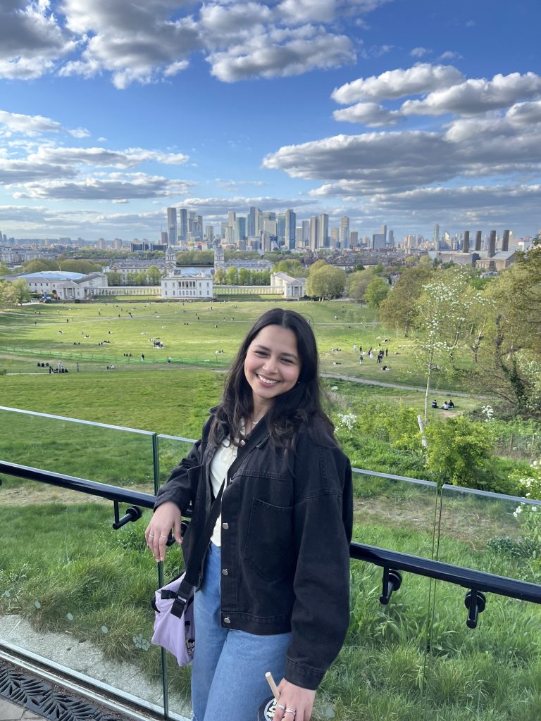A smiling woman stands in front of a glass railing overlooking a green park with scattered people, city buildings, and a partly cloudy sky in the background—capturing one of those memorable student stories at Vita Student.