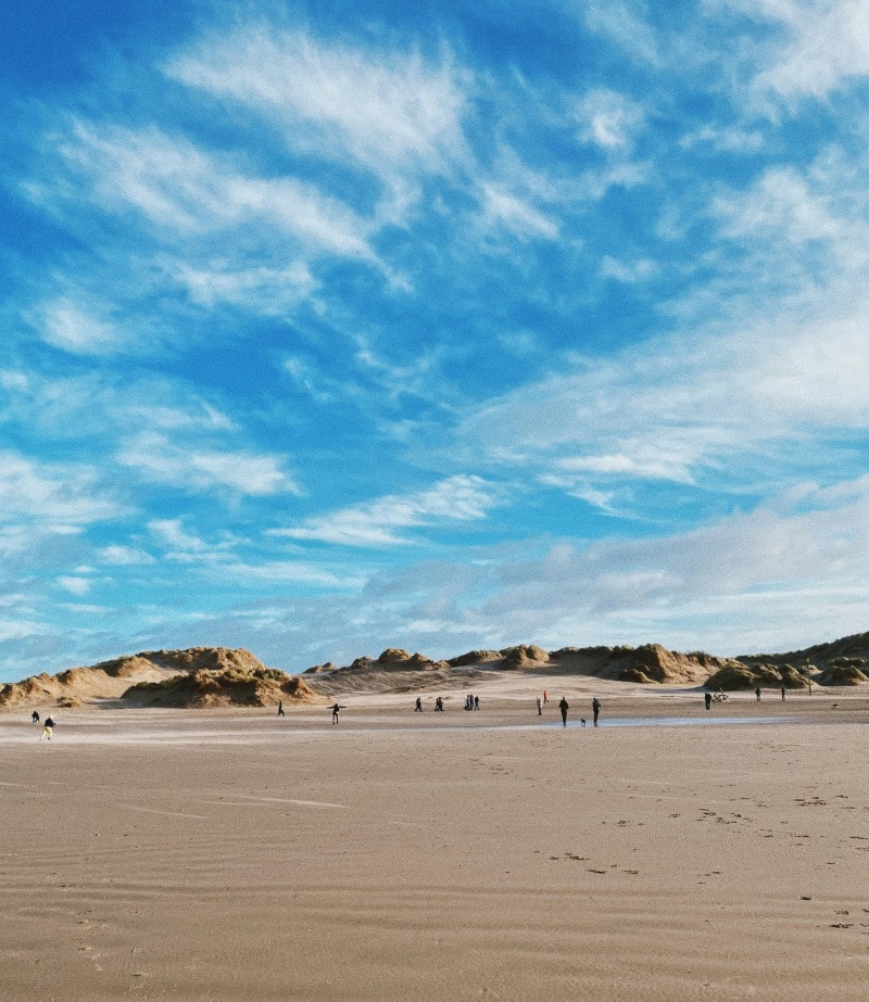 People, including students on day trips from Manchester, walk and stand on a wide sandy beach with sand dunes in the distance under a bright, partly cloudy blue sky.