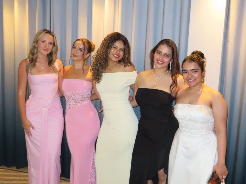 Five women stand together, smiling, dressed in elegant evening gowns in pastel pink, cream, black, and white, in front of a curtain—capturing the spirit of student stories as friends from diverse backgrounds gather to celebrate their journey as Indian students in the UK.