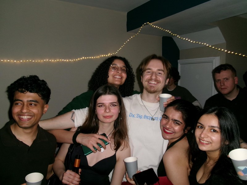 A group of seven young adults, including an Indian student, smiling and posing together at an indoor party, holding cups. String lights are hung on the wall in the background, capturing one of many memorable student stories about adapting to the UK.