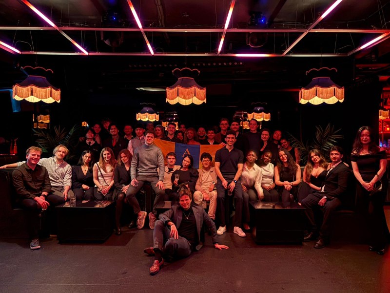 A large group of people, including Indian students, pose together in a dimly lit room with red and pink lights, sitting and standing on sofas. Vintage lamps hang from the ceiling, creating a warm and festive atmosphere that echoes the student stories of adapting to the UK.