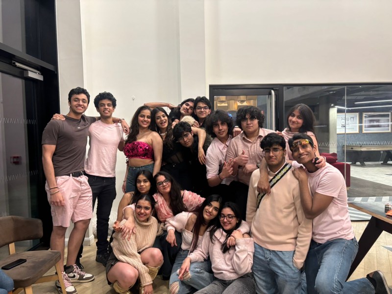 A group of young adults, some standing and some sitting, pose closely together indoors. Most are wearing shades of pink and beige, smiling and appearing happy in a casual, well-lit room with glass walls—a perfect setting to capture student stories of Indian students adapting to the UK and building new friendships.