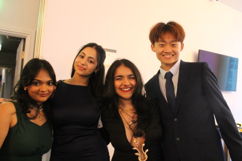Four young adults dressed formally stand together indoors, smiling at the camera—perhaps sharing their own student stories of adapting to the UK. There is a television on the wall behind them, highlighting this moment in the journey of an Indian student abroad.