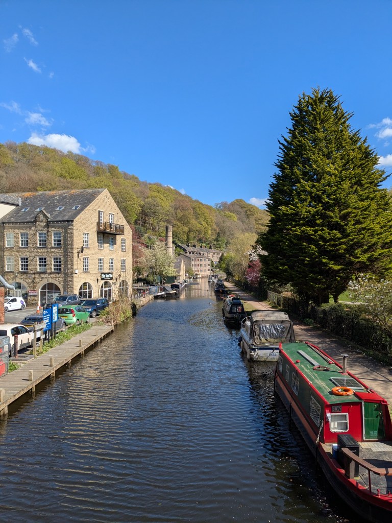 A canal lined with narrowboats and historic stone buildings, bordered by trees and hills under a blue sky with scattered clouds—an ideal destination for day trips for students in Manchester seeking picturesque scenery and a touch of history.