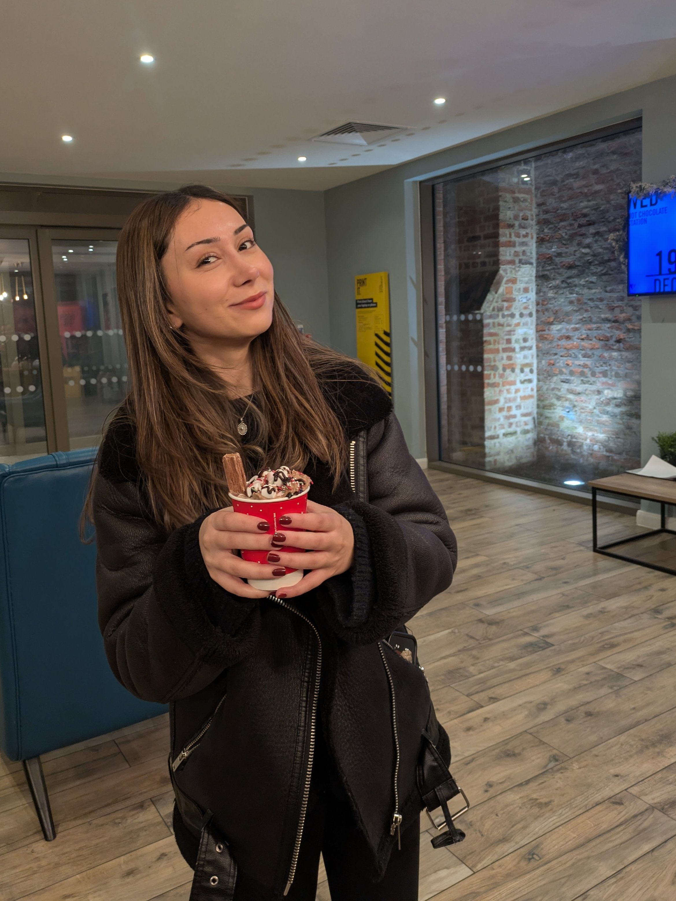 A young woman with long brown hair, wearing a black jacket, stands indoors at York, holding a cup of dessert with whipped cream and chocolate. She is smiling, and the room has wooden floors and modern decor—a true moment from Student Stories.