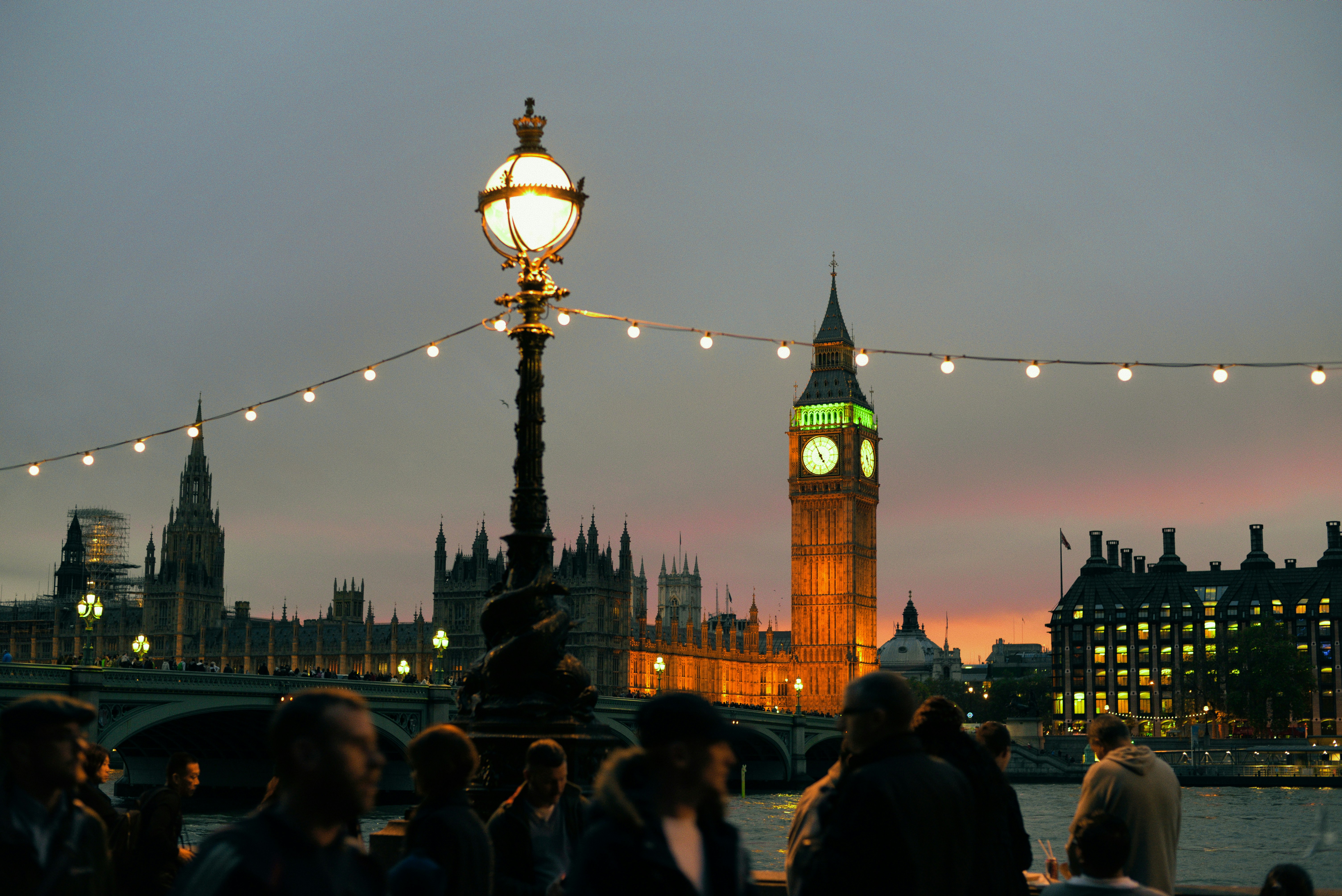 People gather by the Thames at dusk, with the illuminated Big Ben and Houses of Parliament in the background, string lights hanging overhead—a picturesque moment in the heart of the UK, recently featured in the QS Best Student Cities 2027 rankings.