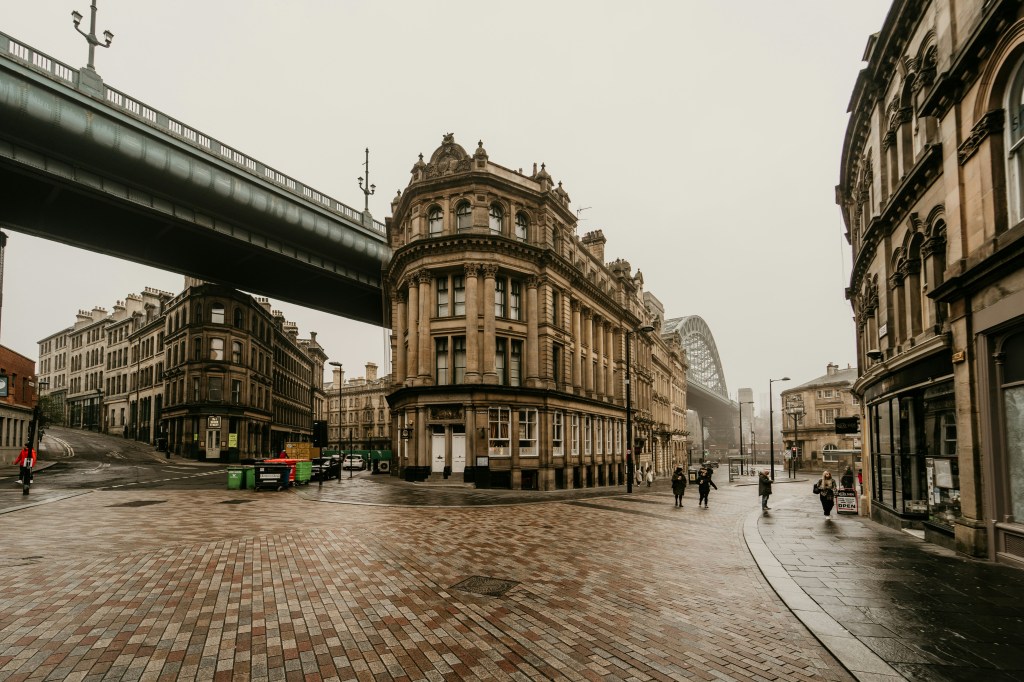 A quiet, overcast city street with historic stone buildings sits near Leazes Park. A curved corner building anchors the view, with an elevated bridge overhead. Few pedestrians, possibly students near Leazes Park, walk the wet cobblestone pavement.