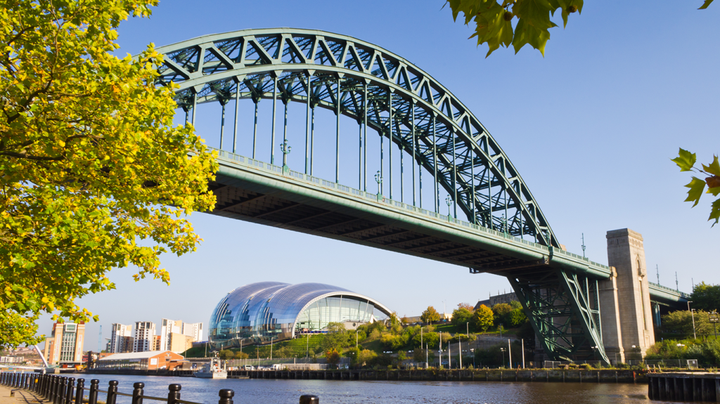 A large steel arch bridge spans over a river with modern glass buildings and green trees on the bank below, under a clear blue sky—perfect for students near Leazes Park seeking things to do or scenic views.