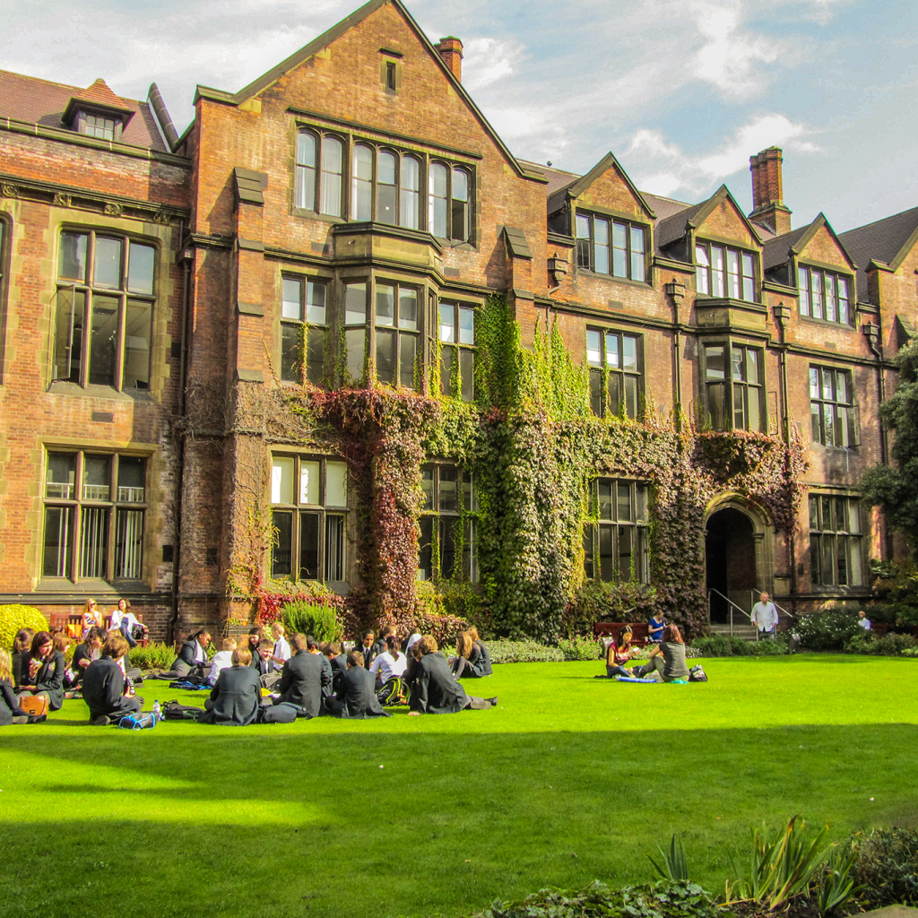 Students near Leazes Park sit in groups on a bright green lawn in front of a large, historic brick building with ivy climbing its walls and tall windows on a sunny day, enjoying one of the many things to do in this scenic area.