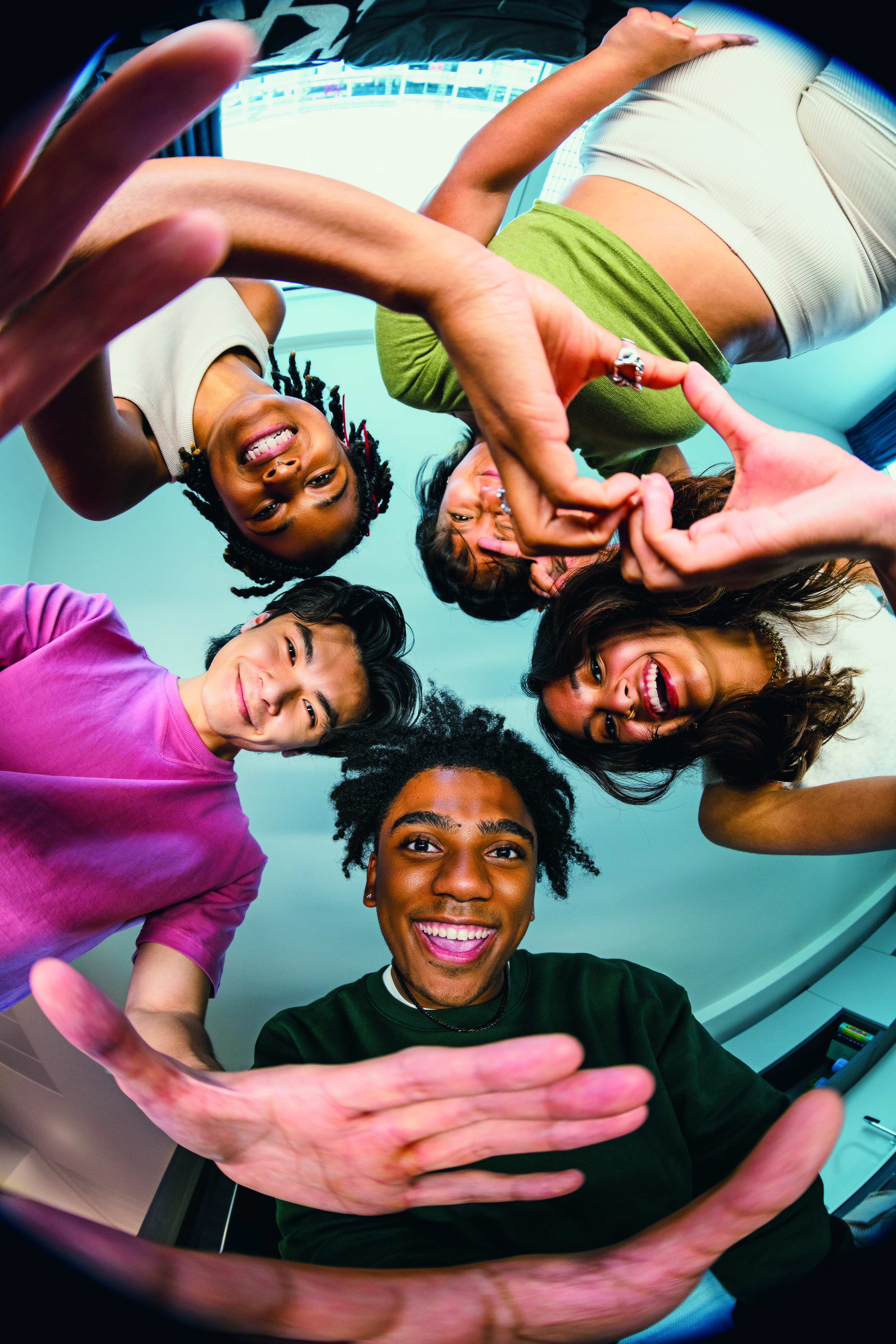 Five smiling young people stand in a circle, looking down at the camera, some making heart shapes with their hands. Captured with a fisheye lens for a playful, distorted effect, this vibrant group radiates positivity—embodying the spirit of HealthHero and celebrating friendship and well-being.