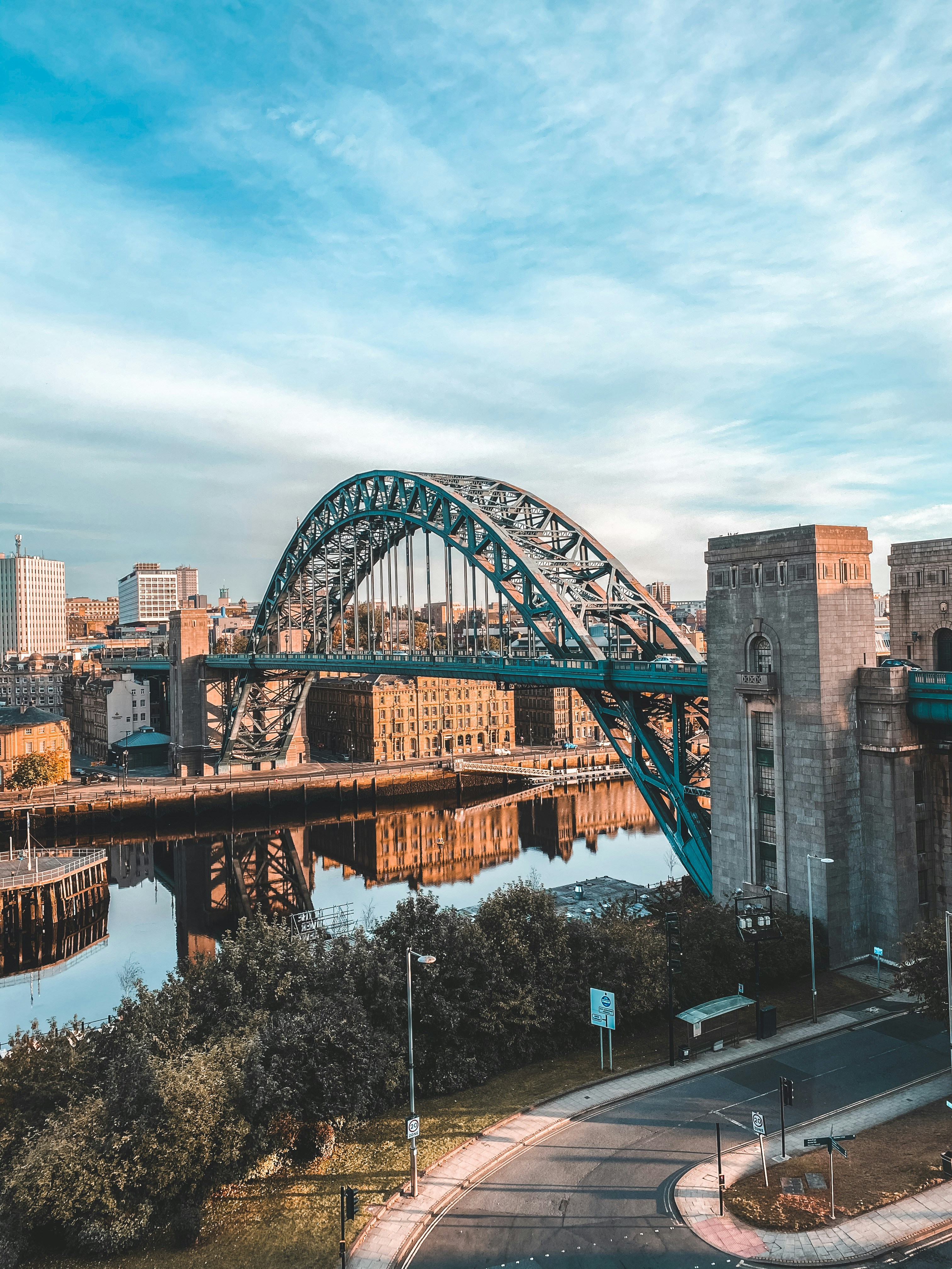 A large steel arched bridge spans a river with city buildings in the background, under a blue sky with light clouds. Students near Leazes Park can enjoy views of trees and a road in the foreground.