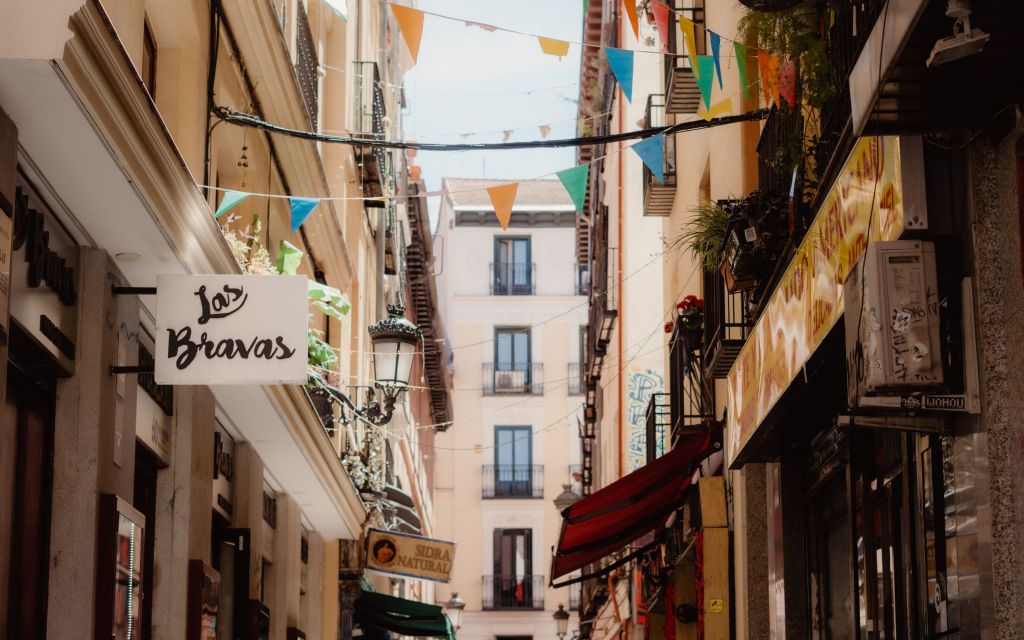 Narrow European street with colorful flags overhead, restaurant signs, balconies with flowers, and tall buildings lining both sides, illuminated by daylight—a classic scene capturing the vibrant atmosphere that makes discovering student activities in Madrid so appealing.
