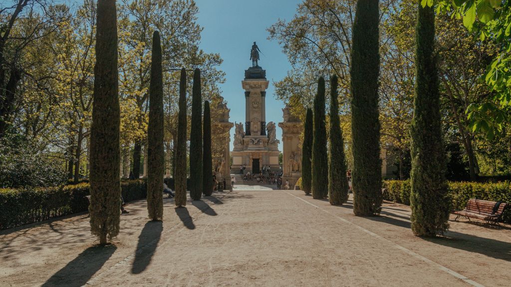 Wide pathway lined with tall cypress trees leads to a monument with a statue on top, surrounded by greenery and people gathered near the structure on a sunny day—a perfect spot for student activities Madrid offers, and one of the many things to do in Madrid for students looking to enjoy outdoor spaces.