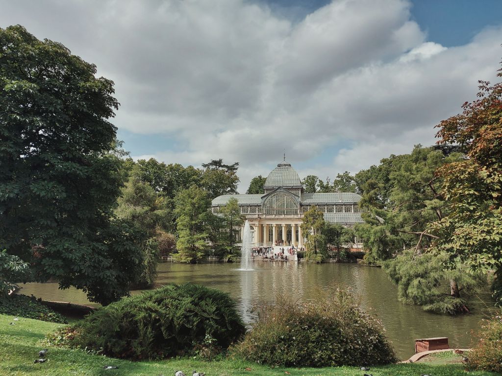 A glass palace sits by a pond, surrounded by lush green trees under a partly cloudy sky. People gather at the building’s entrance, and the water reflects the structure and foliage—a must-see spot often featured on lists of things to do in Madrid, especially popular among student activities Madrid has to offer.