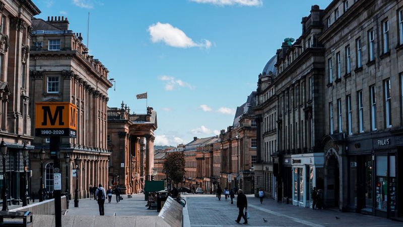 A city street lined with historic stone buildings, people walking along the sidewalks, and a blue sky with a few clouds overhead. A yellow sign with the letter "M" is visible on the left, marking one of the best areas Newcastle has to offer for student accommodation Newcastle locals recommend.