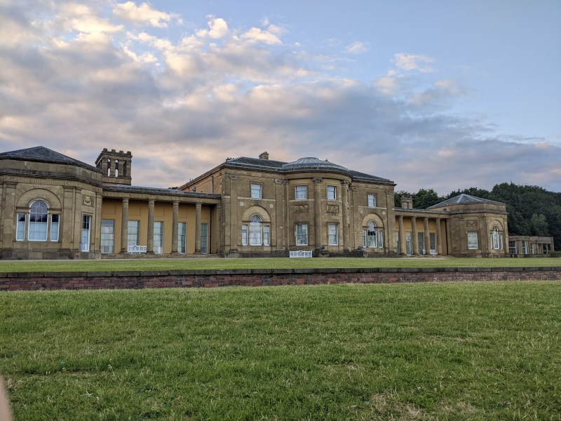 Large historic building with arched windows and columns, set against a cloudy sky in Manchester. The grass lawn in front is well-kept, and trees are visible behind the structure, making it an ideal spot for students on day trips to relax and explore.