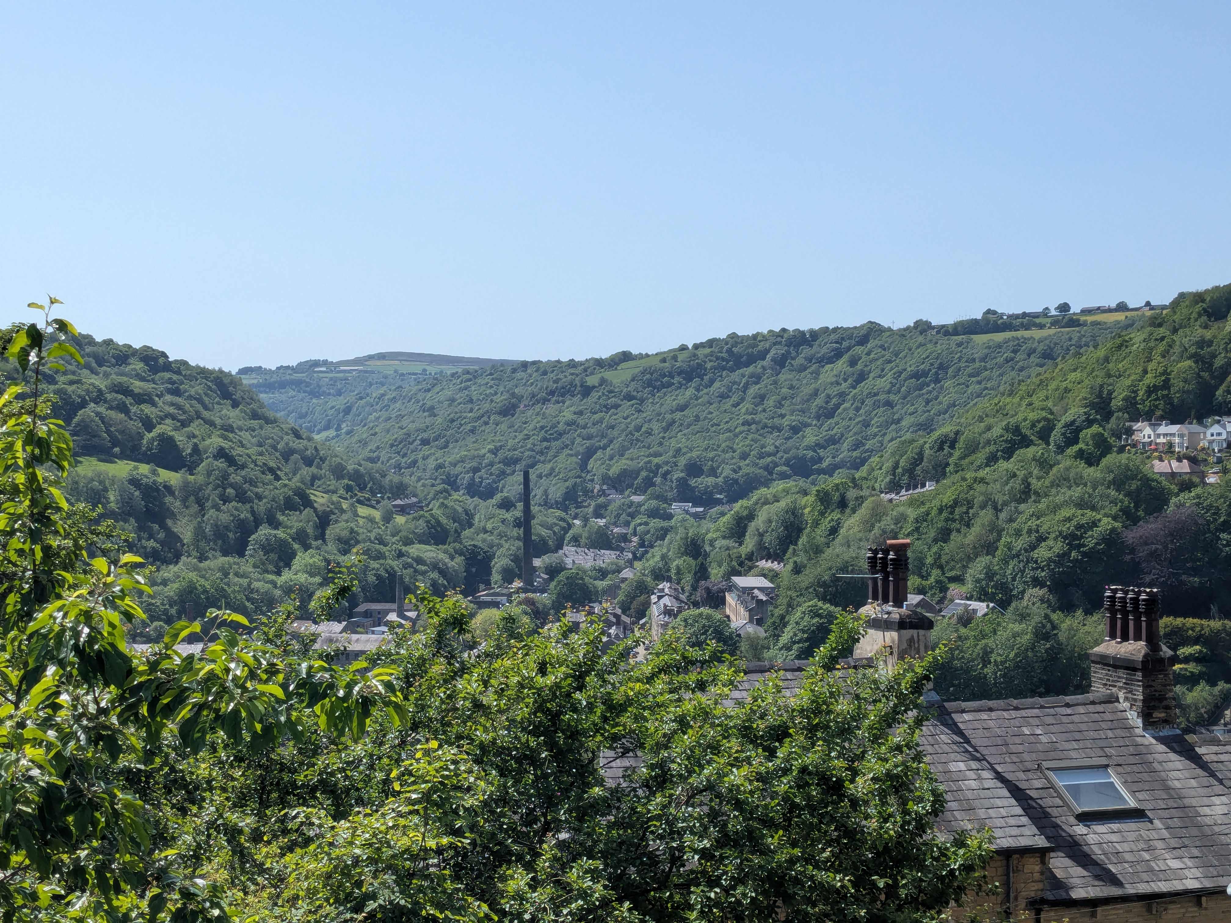 A lush green valley with dense trees, rooftops with chimneys in the foreground, and hills rising in the background under a clear blue sky—an idyllic spot often chosen by students from Manchester for day trips.