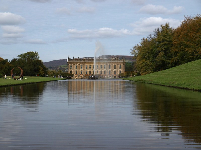 A grand historic mansion stands beyond a large, reflective pond with a tall water fountain at its center, flanked by green lawns and autumn trees under a partly cloudy sky—making it an ideal spot for students from Manchester seeking memorable day trips.